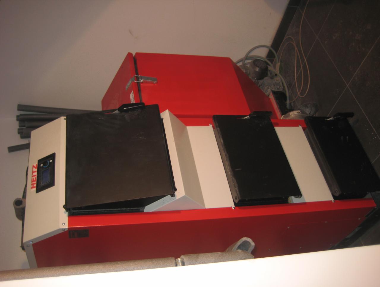 Czerwono-biały piec na paliwo stałe marki Heiz z czarnymi drzwiczkami, widoczny w rogu pomieszczenia z ciemną podłogą i przewodami. Ujęcie z góry.