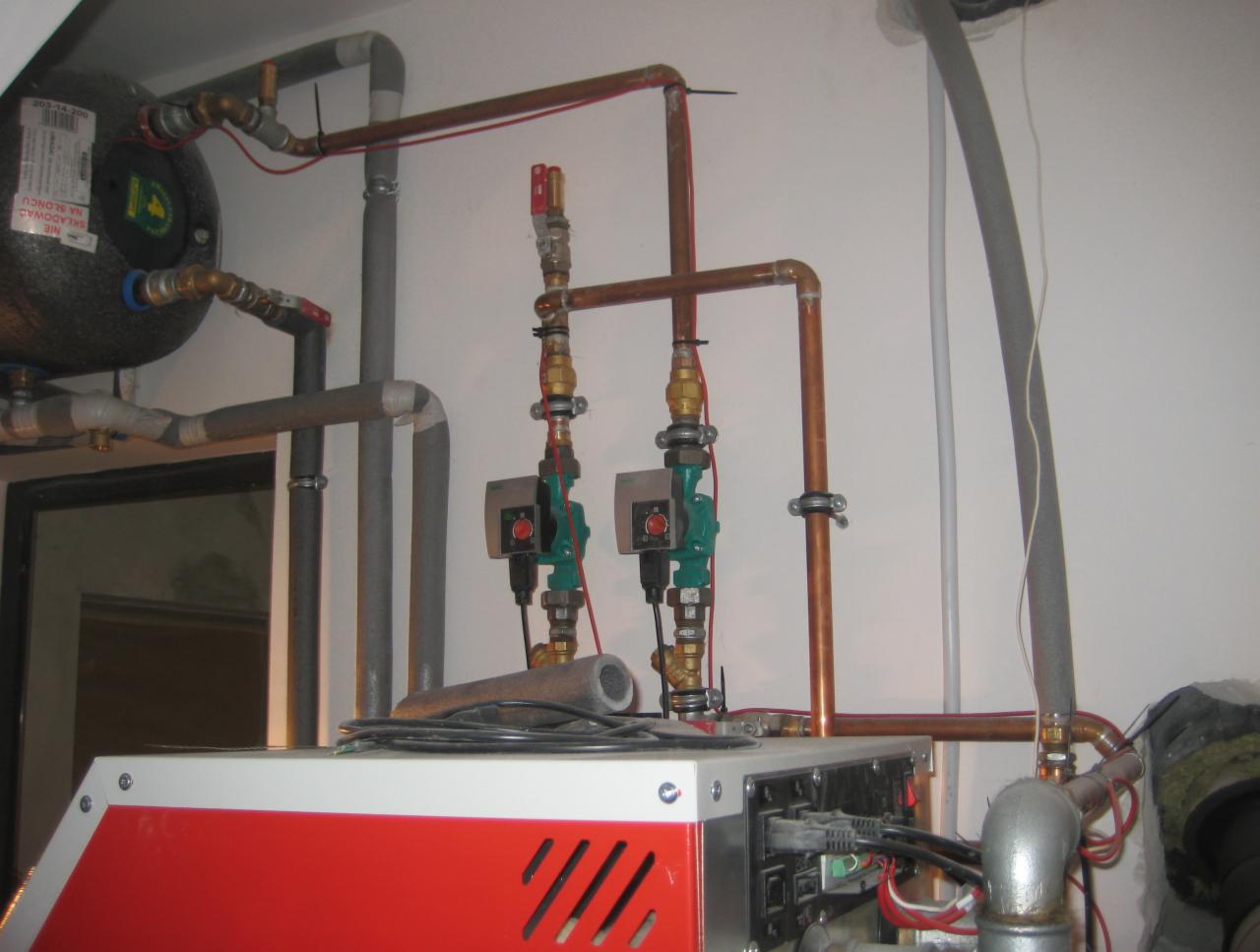Instalacja grzewcza z czerwonym kotłem i miedzianymi rurami. Widoczne zawory, pompy obiegowe i zbiornik. Szare rury izolacyjne. Białe ściany. Ujęcie z bliska.