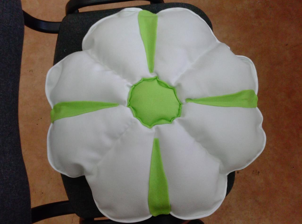 Dekoracyjna poduszka w kształcie kwiatu z białymi płatkami i zielonym środkiem, leżąca na krześle. Widoczne przeszycia i detale wykończenia.