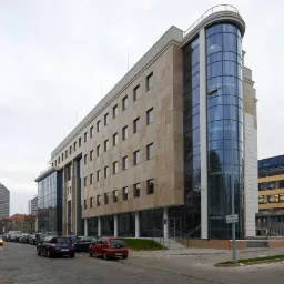 Nowoczesny budynek biurowy z kamienną elewacją i przeszkloną, zaokrągloną częścią narożną, widoczny z poziomu ulicy z zaparkowanymi samochodami.