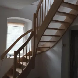 Jasne, drewniane schody kręte w minimalistycznym wnętrzu z oknem łukowym. Widoczne detale balustrady i stopnie, oświetlone naturalnym światłem.