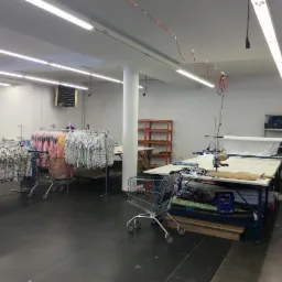 Jasne wnętrze szwalni z wózkami pełnymi odzieży, stołami do krojenia i maszynami do szycia. Widoczne materiały i gotowe ubrania. Oświetlenie jarzeniowe.