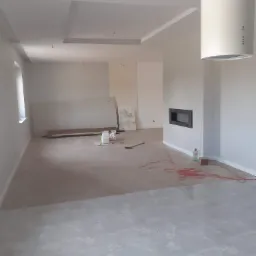 Wnętrze w trakcie remontu: białe ściany, marmurowe płytki podłogowe, zabudowany kominek, podwieszany sufit z oświetleniem punktowym, widoczne materiały budowlane i przewody na podłodze.