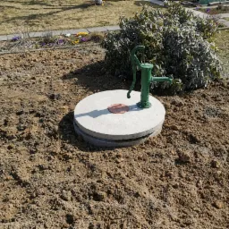 Zielona pompa ręczna zamontowana na betonowym kręgu studni, otoczona świeżo przekopaną ziemią, z krzewami i kwiatami w tle.