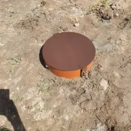 Brązowa, okrągła pokrywa studni rewizyjnej z zamkiem, umieszczona w ziemi, widoczny cień osoby robiącej zdjęcie.