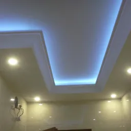 Podwieszany sufit z oświetleniem LED w kształcie litery V, punktowe lampy halogenowe, beżowe ściany wykończone płytkami. Dekoracyjne oświetlenie sufitowe.