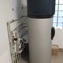 Szary piec z podłączonymi rurami w narożniku pomieszczenia z oknem z luksferami, widoczny fragment instalacji hydraulicznej i mały zbiornik wyrównawczy.