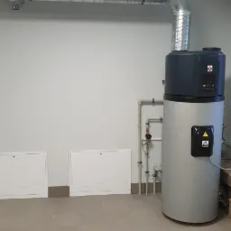 Nowoczesny, srebrny piec grzewczy z szarą górną częścią, podłączony do rury wentylacyjnej w jasnym pomieszczeniu z kafelkami na podłodze i białymi szafkami rewizyjnymi na ścianie.