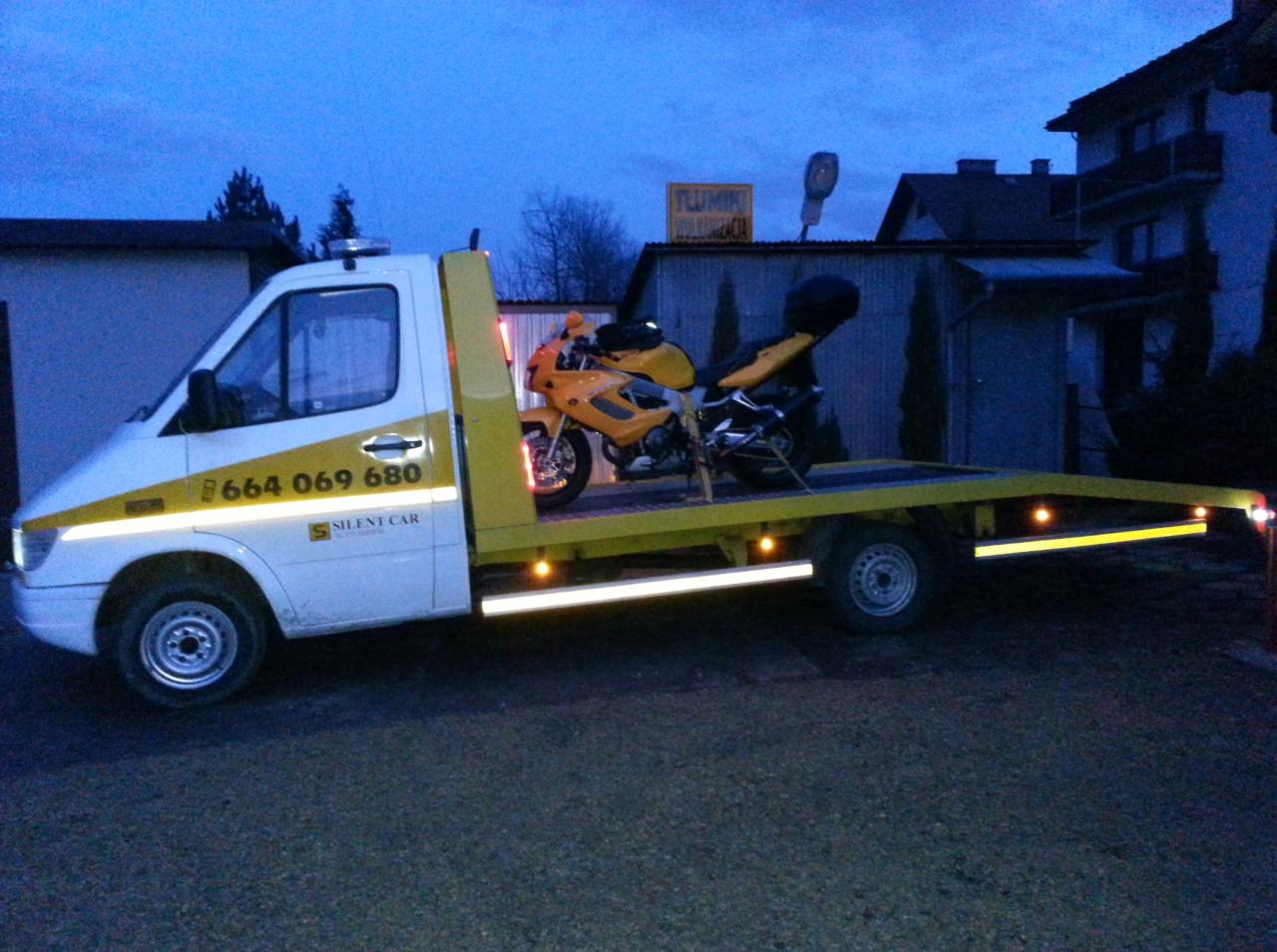 Żółty motocykl na platformie białej lawety z numerem telefonu i logo 'Silent Car' na tle budynków w wieczornym oświetleniu.