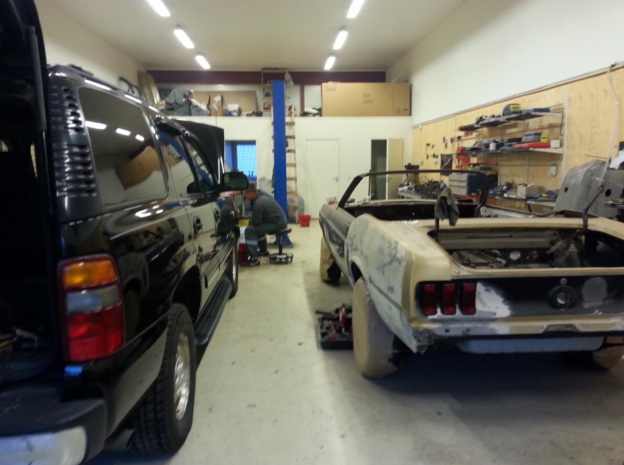 Warsztat samochodowy: czarny SUV z otwartą maską i odrestaurowywany kabriolet Mustang. Mechanik przy SUV-ie, narzędzia na półkach. Specyficzne oświetlenie LED.