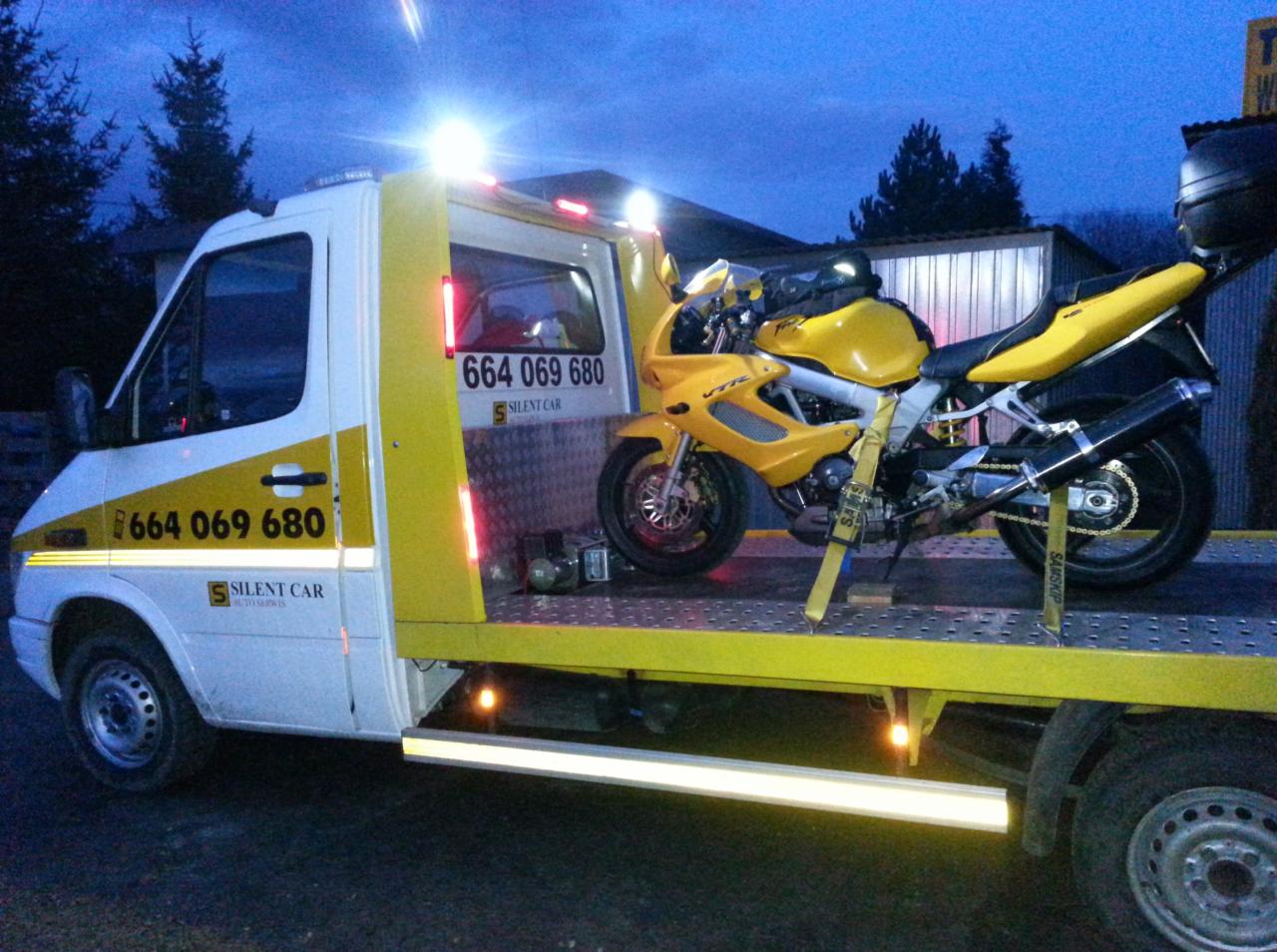 Żółty motocykl VTR na lawecie Silent Car z numerem 664 069 680, zabezpieczony pasami transportowymi. Ujęcie o zmierzchu, widoczne światła lawety.