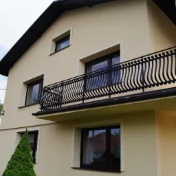 Elewacja domu po remoncie z beżową fasadą, ciemnobrązową stolarką okienną i balkonem z ozdobną, metalową balustradą. Uwagę przykuwa równa powierzchnia ścian.