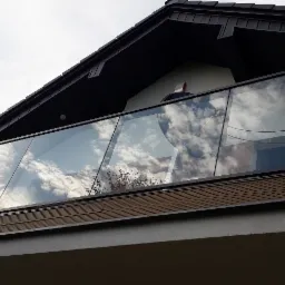 Nowoczesna szklana balustrada balkonowa z czarnymi profilami, odbijająca chmury, zamontowana na jasnym budynku z ciemnym dachem.