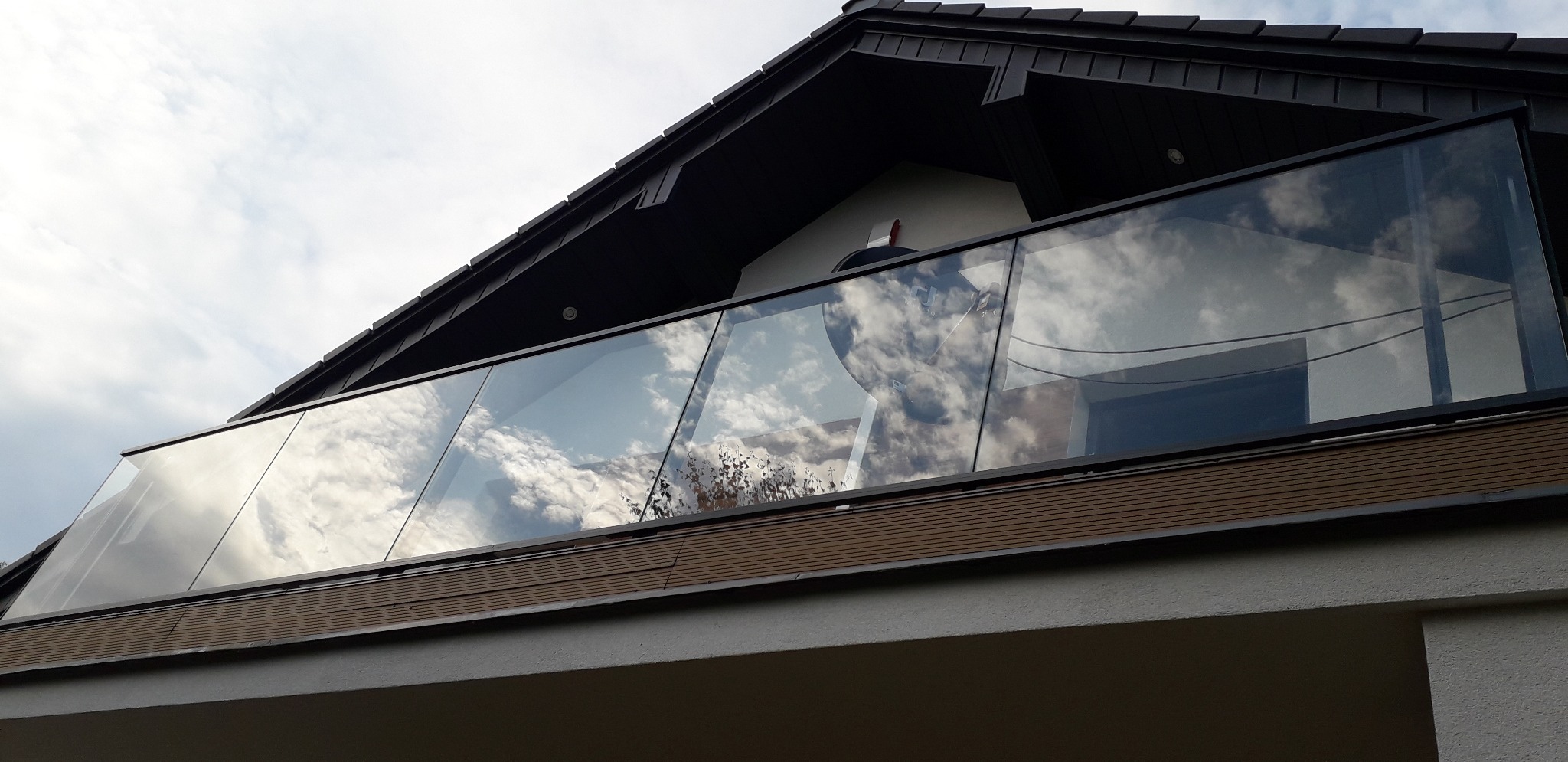 Nowoczesna szklana balustrada balkonowa z czarnymi profilami, odbijająca chmury, zamontowana na jasnym budynku z ciemnym dachem.