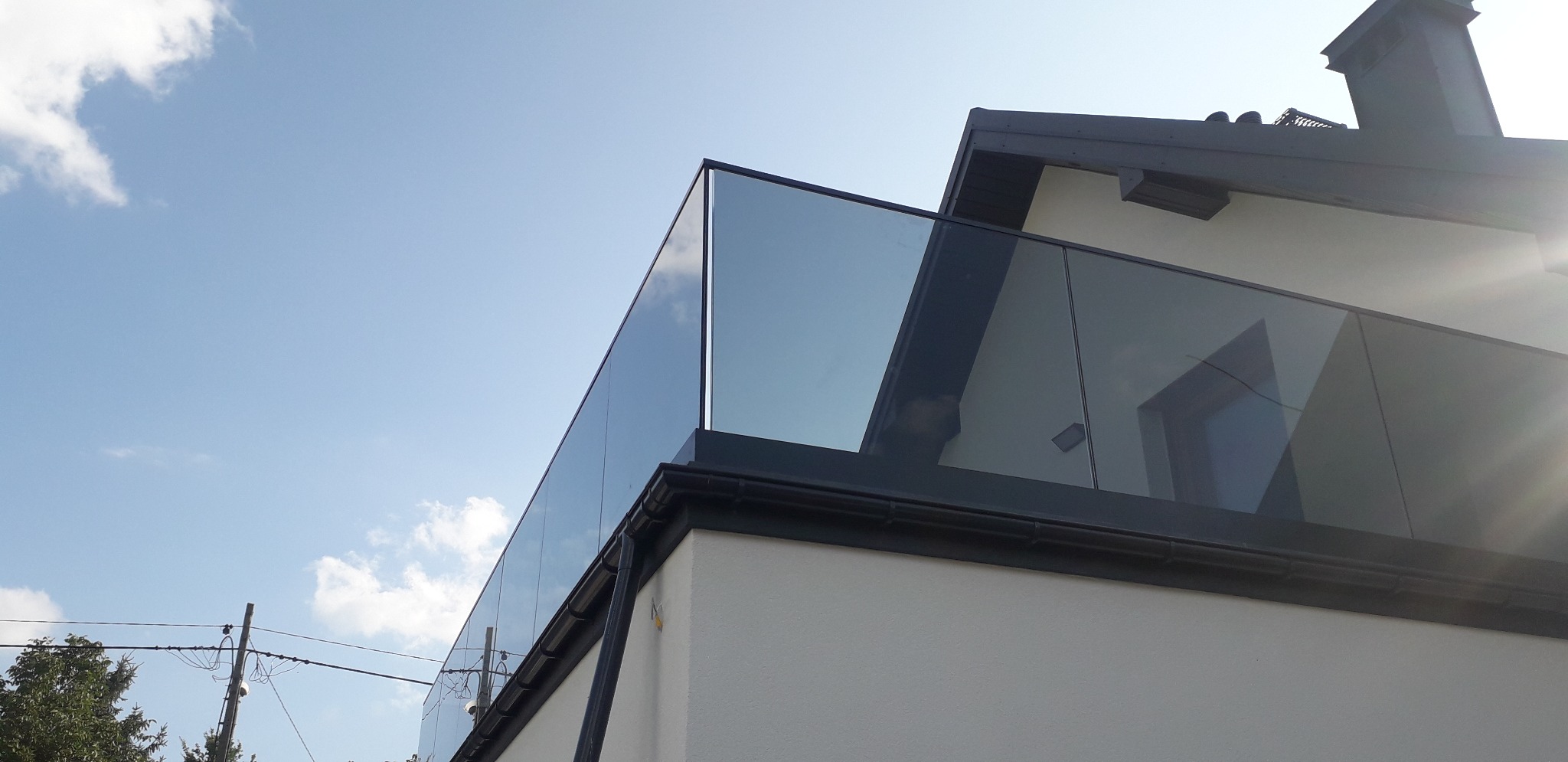 Nowoczesna szklana balustrada balkonowa z czarnymi profilami, widok z dołu na tle błękitnego nieba z chmurami.