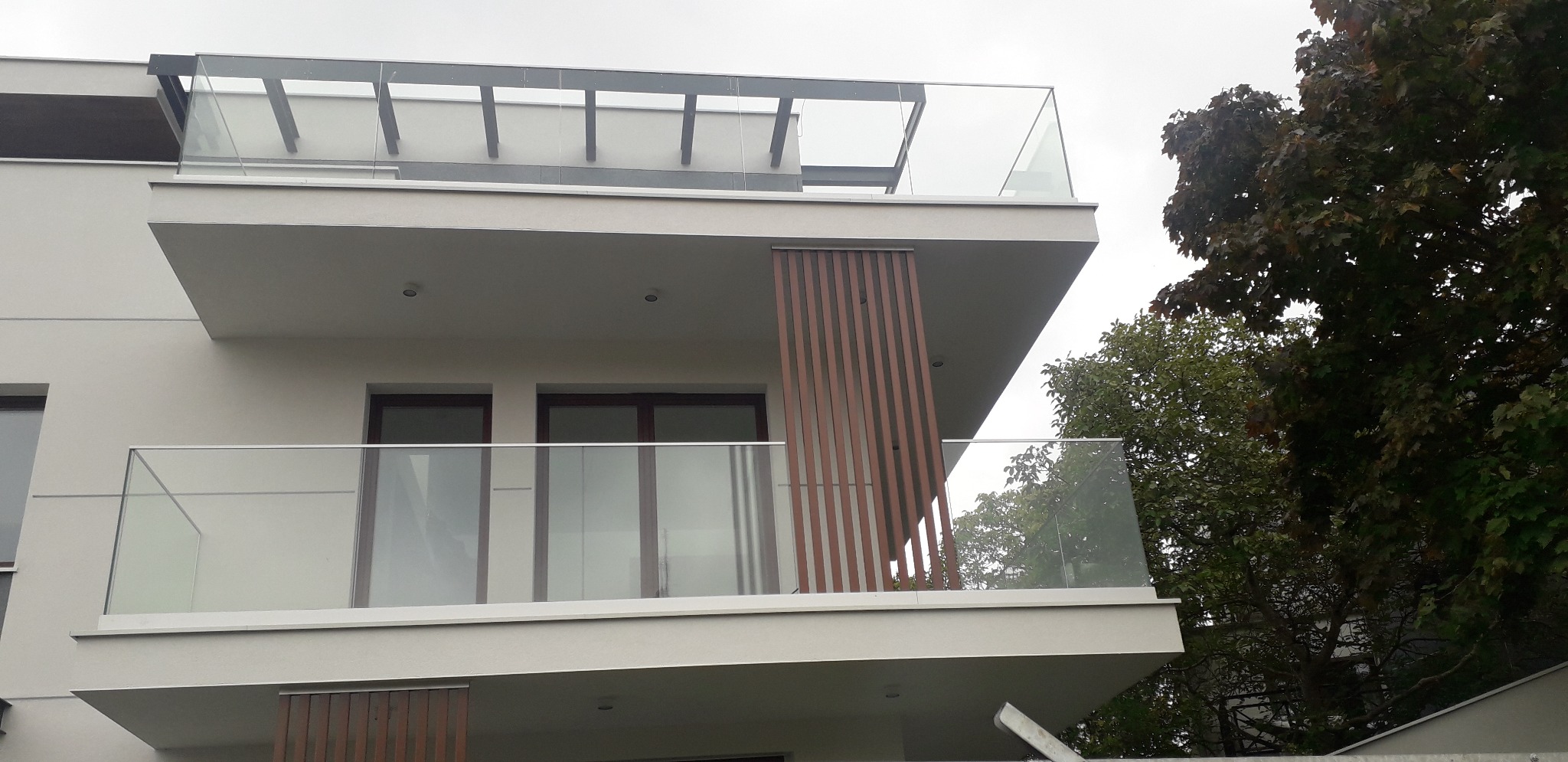 Nowoczesny budynek z balkonami zabezpieczonymi przezroczystymi taflami szkła, widoczne pionowe, drewnopodobne panele dekoracyjne