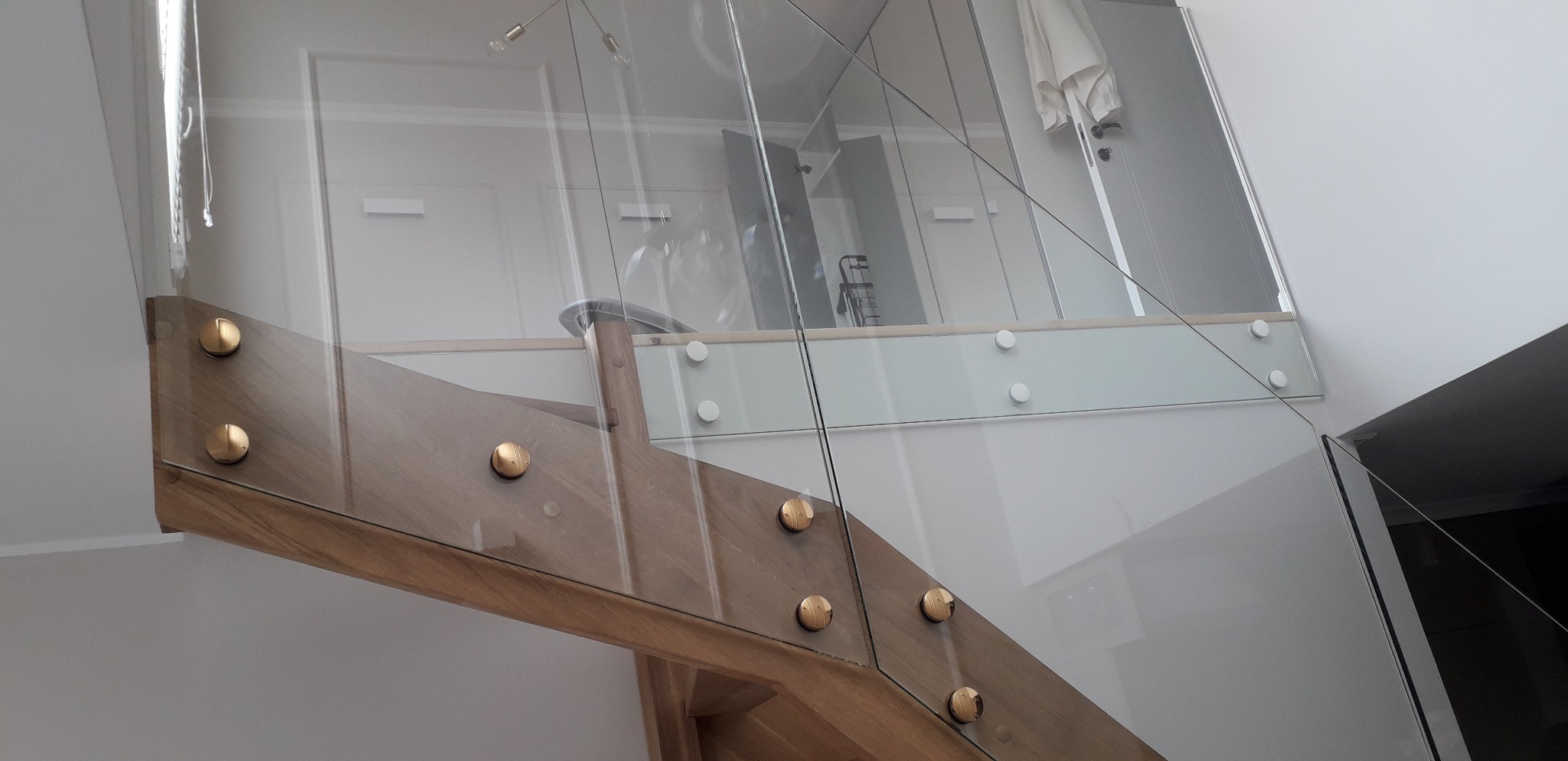 Szklana balustrada schodowa z drewnianymi elementami mocującymi i złotymi śrubami, widok z dołu na górny poziom.