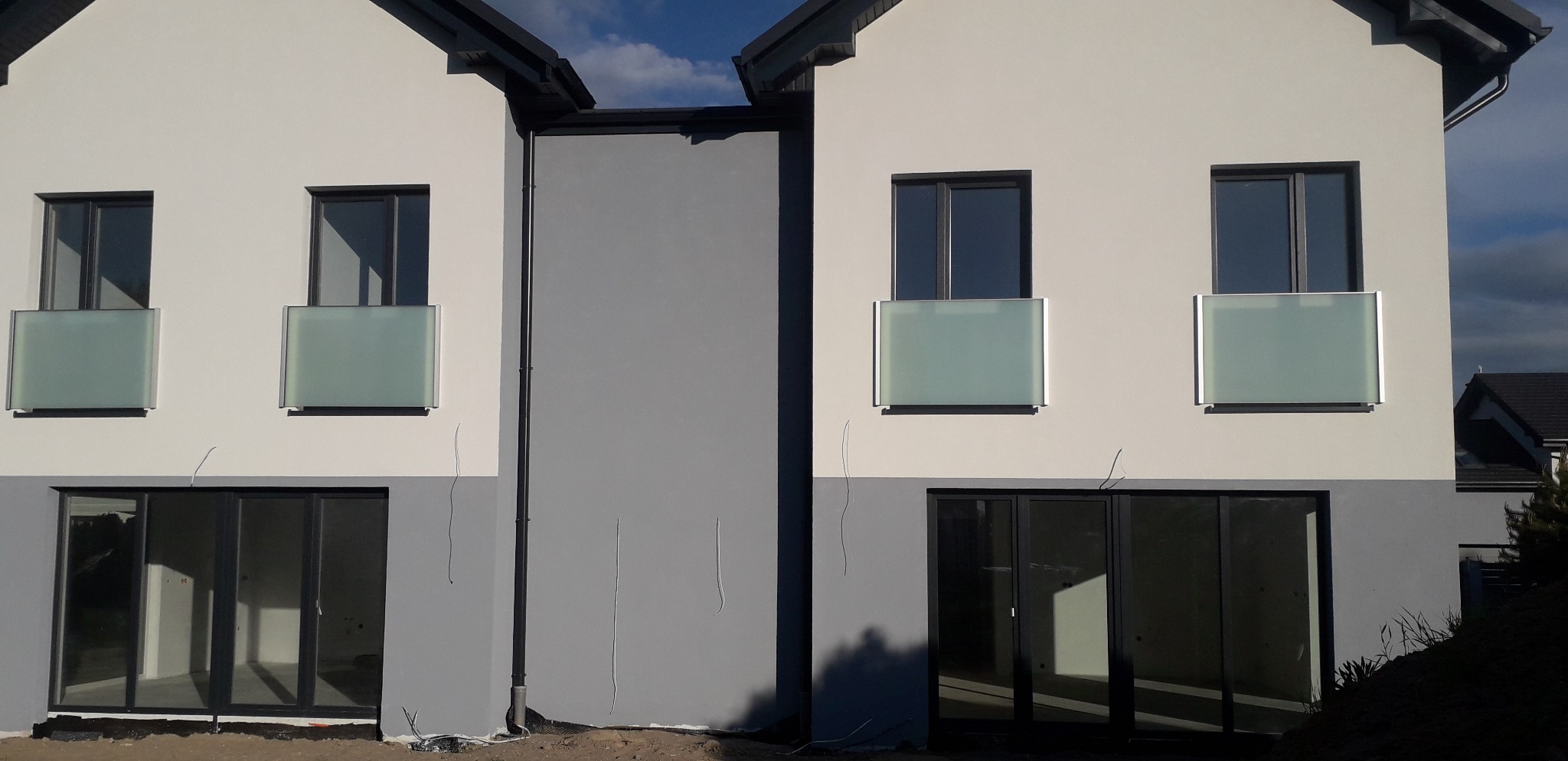 Dwa bliźniacze domy w trakcie wykończenia, z szklanymi barierkami balkonowymi zamontowanymi na pierwszym piętrze, widoczne niedokończone otoczenie i instalacje.