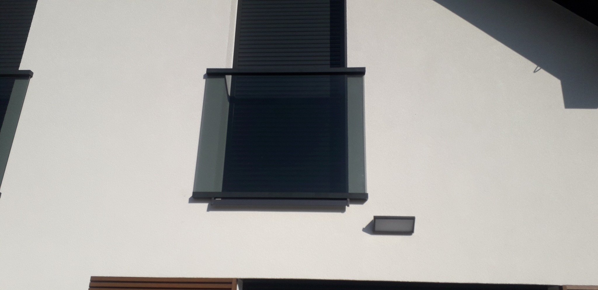 Nowoczesna przeszklona balustrada balkonowa z szarymi ramami, zamontowana na białej fasadzie budynku z roletami zewnętrznymi.