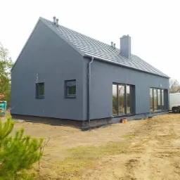 Nowoczesny dom jednorodzinny z szarą elewacją i ciemnym dachem, widok z zewnątrz, teren wok&oacute;ł w trakcie zagospodarowania, widoczny fragment białego samochodu dostawczego.