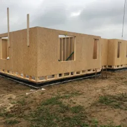 Wznoszenie ścian parteru domu z drewnianych paneli OSB na fundamencie, widoczne otwory okienne i drzwiowe, częściowo wykończona podstawa, na zewnątrz teren budowy.