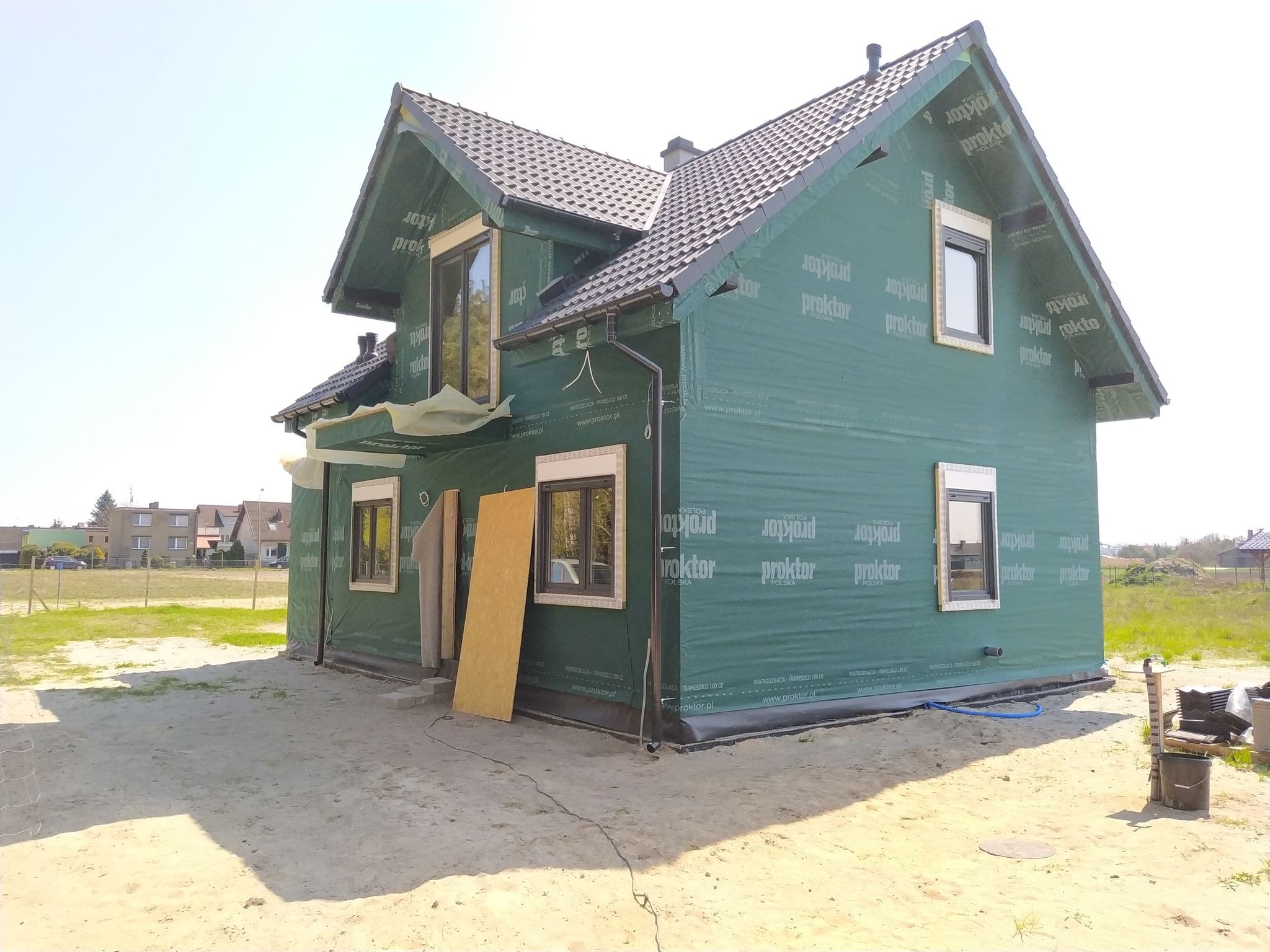 Surowy stan domu z zieloną membraną wiatroizolacyjną, widoczne okna z brązowymi ramami, płyta OSB oparta o ścianę, teren wokół domu piaszczysty.