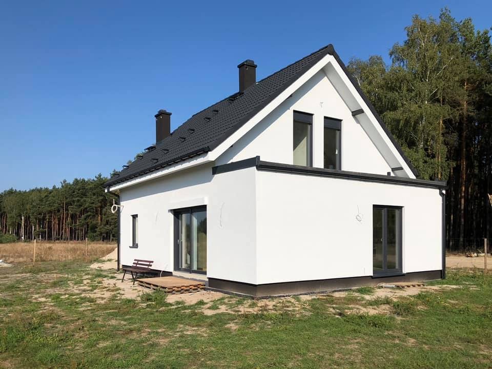 Nowoczesny, biały dom z czarnym dachem i oknami, z małą ławeczką na drewnianym podeście przed wejściem, na tle lasu i błękitnego nieba.