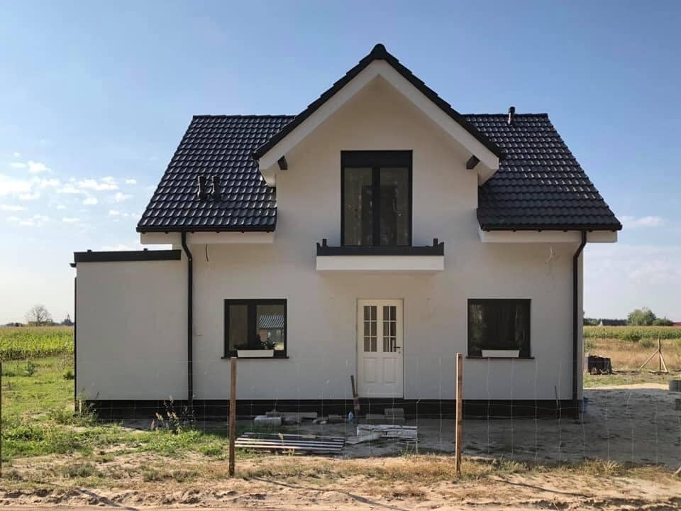 Surowy, biały dom jednorodzinny z ciemną dachówką i czarnymi ramami okiennymi, w trakcie budowy, z widocznym ogrodzeniem z siatki i polami w tle.