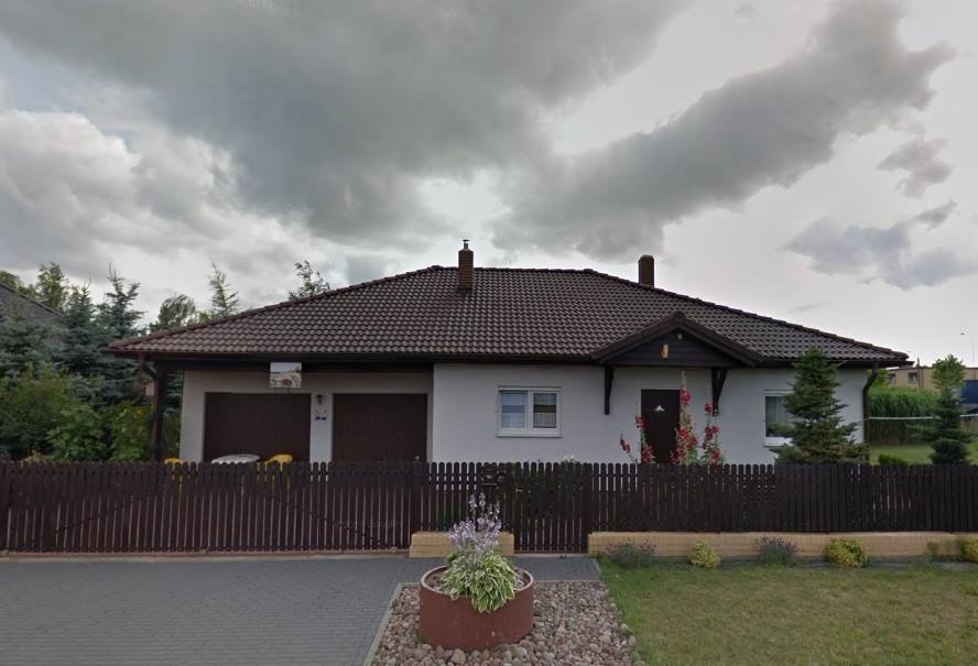 Parterowy dom z brązowym dachem krytym dachówką, szarymi ścianami, brązową stolarką okienną i drzwiową oraz drewnianym ogrodzeniem, widziany z przodu, z elementami zagospodarowania terenu przed domem.