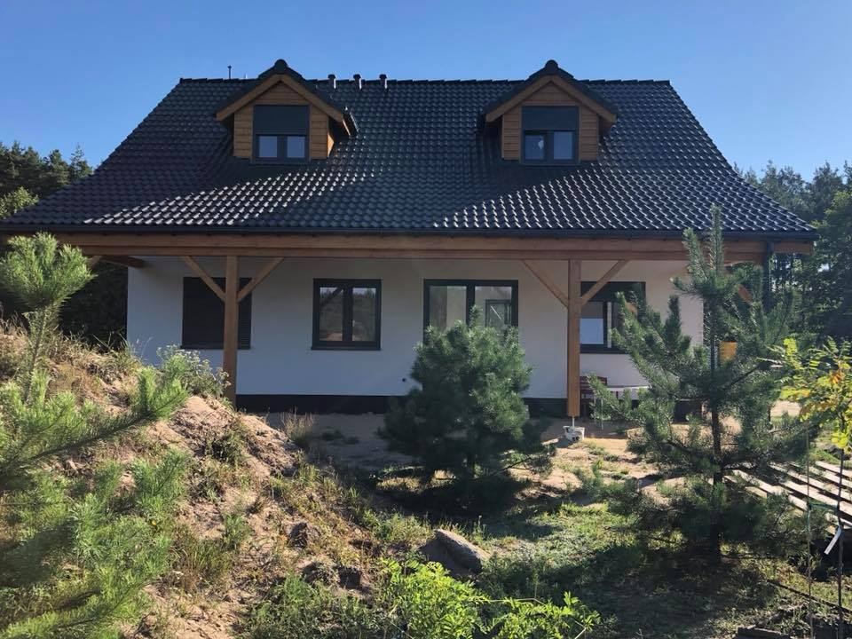 Gotowy dom z białymi ścianami, ciemną dachówką i drewnianą konstrukcją werandy, otoczony młodymi sosnami na piaszczystym terenie.