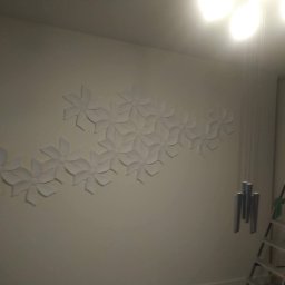 Greg-Bud Usługi Remontowo Budowlane - Ściana z dekoracyjnymi, białymi elementami w kształcie kwiatów, drabina aluminiowa i metalowe dzwonki w tle.