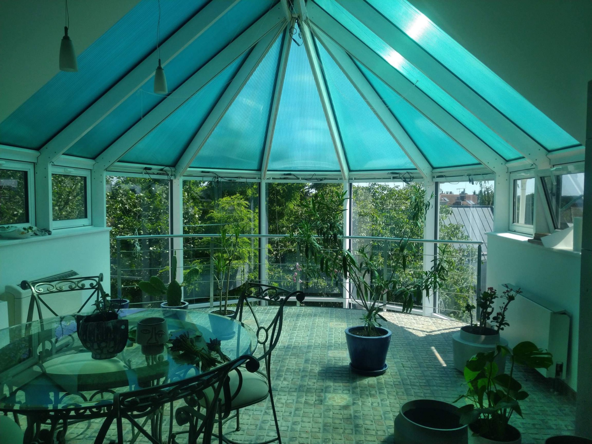 Zadaszony taras w Warszawie z przeszklonym dachem w odcieniach turkusu, szklanym okrągłym stołem i metalowymi krzesłami, otoczony zielenią.