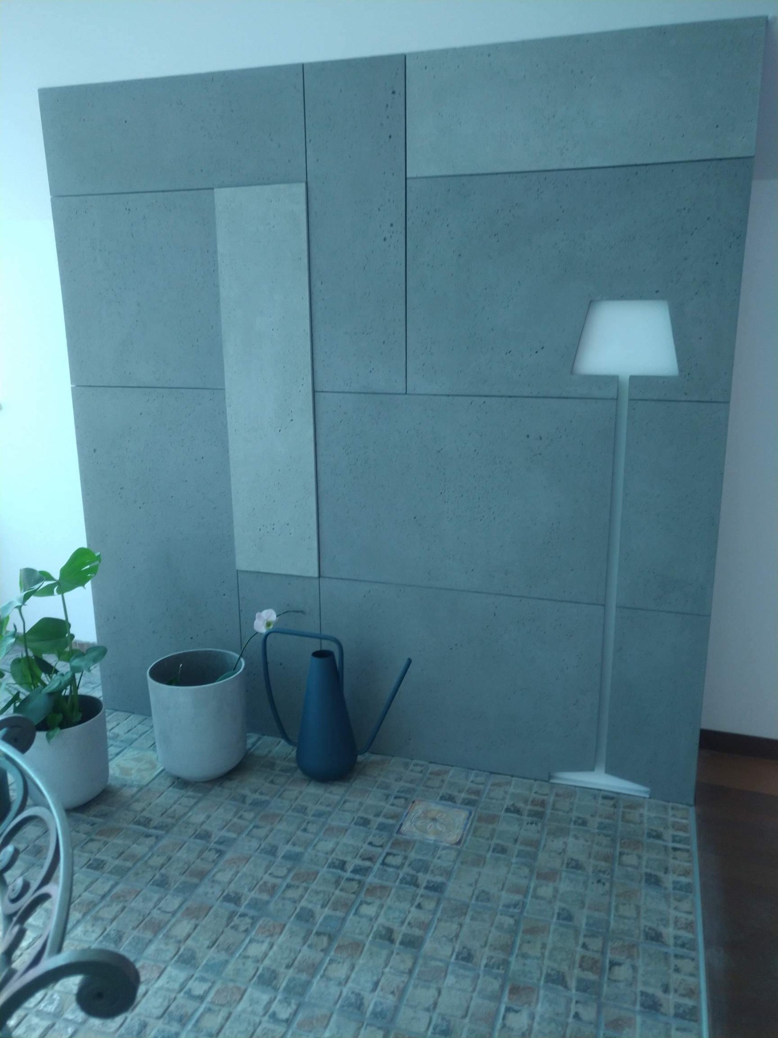 Ściana wykończona panelami z szarego betonu architektonicznego o zróżnicowanych rozmiarach, uzupełniona o minimalistyczną lampę podłogową, roślinę w doniczce i niebieską konewkę na mozaikowej...