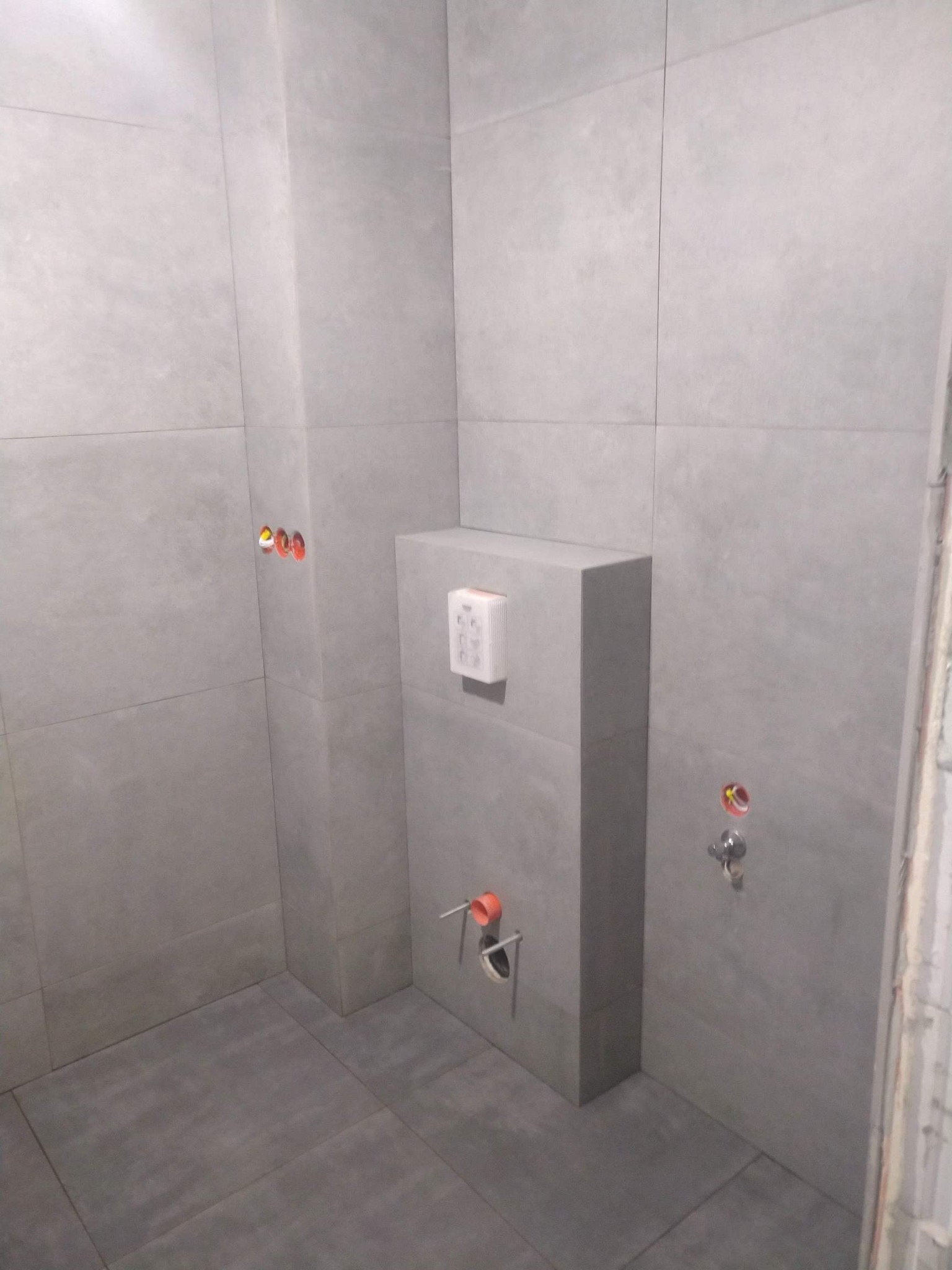 Szara łazienka w trakcie wykańczania, widoczne płytki na ścianach i podłodze, stelaż podtynkowy WC z przyciskiem spłukującym, wystające rury instalacji wodnej i kanalizacyjnej oraz otwory...