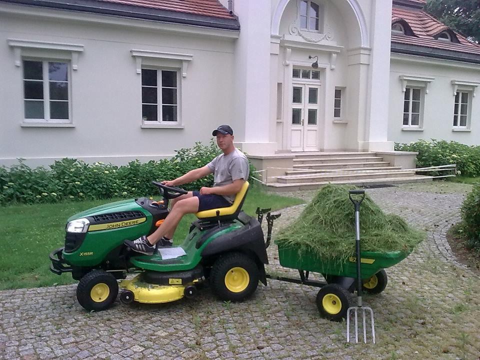 Mężczyzna na kosiarce John Deere X155R z przyczepką pełną trawy na tle okazałego domu. Wózek z trawą i widły ogrodowe.
