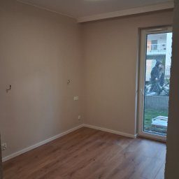 Damian Wilk - Pusty pokój po malowaniu ścian na beżowo i ułożeniu paneli podłogowych z widocznym oknem balkonowym i lampą sufitową.