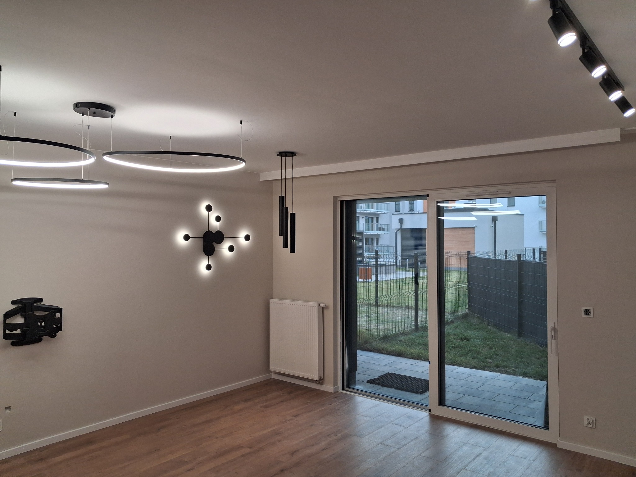 Nowoczesny salon z drewnianą podłogą, oświetleniem LED: okrągłe lampy wiszące, reflektory szynowe, dekoracyjna lampa ścienna w kształcie atomu, oraz czarna abstrakcyjna rzeźba ścienna. Duże...