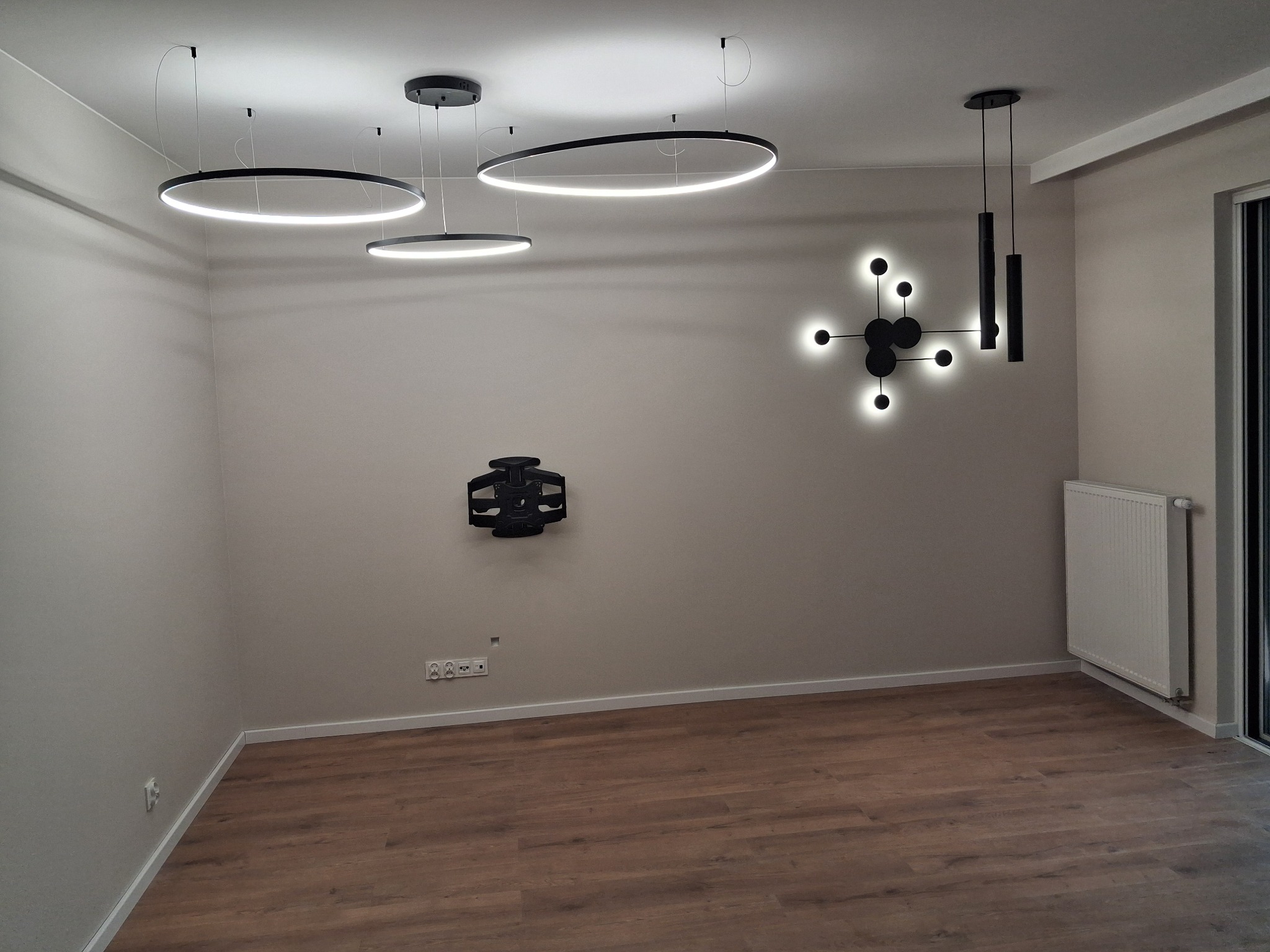 Nowoczesne oświetlenie sufitowe z okrągłych lamp LED i dekoracyjnej lampy ściennej w kształcie atomu, zamontowany uchwyt na telewizor na ścianie, panele podłogowe w odcieniach brązu, grzejnik przy...