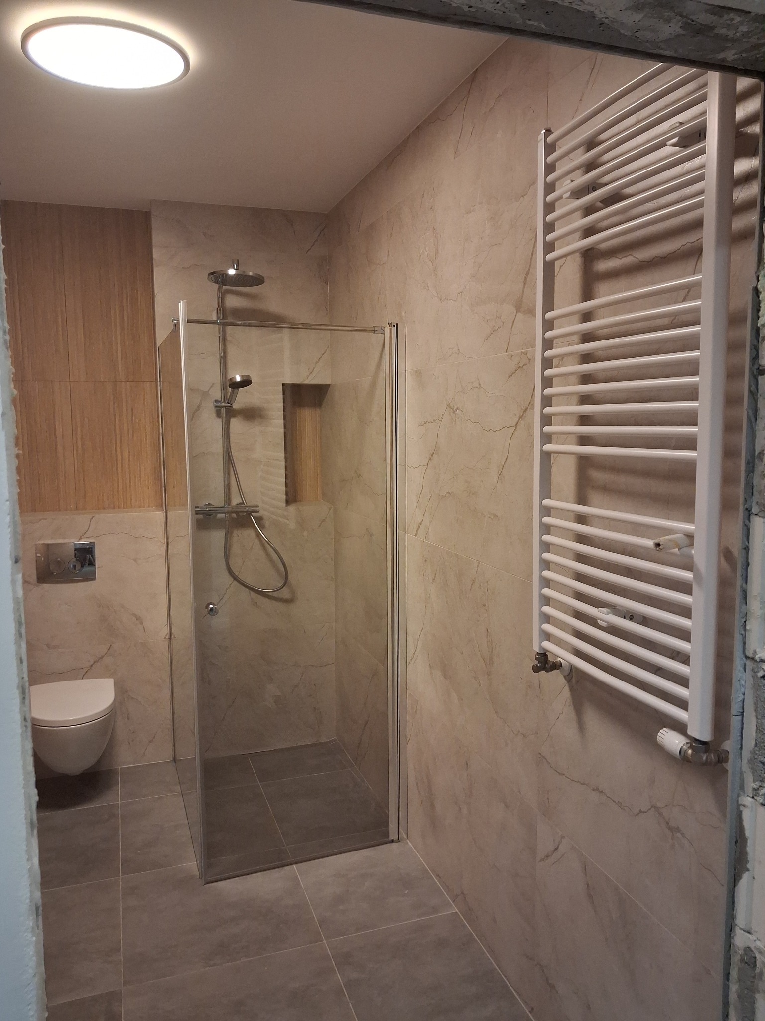 Nowoczesna łazienka z kabiną prysznicową typu walk-in, grzejnikiem drabinkowym i podwieszaną toaletą, wykończona płytkami w odcieniach beżu i szarości.