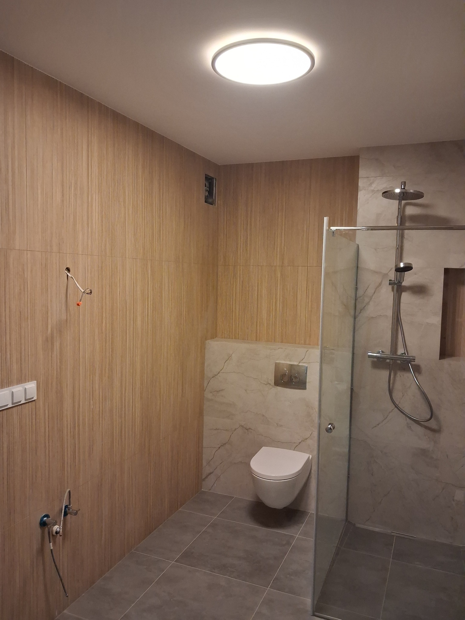Wykończona łazienka z drewnopodobnymi panelami ściennymi, szklaną kabiną prysznicową i podwieszaną toaletą, widoczne niedokończone podłączenia elektryczne i hydrauliczne.