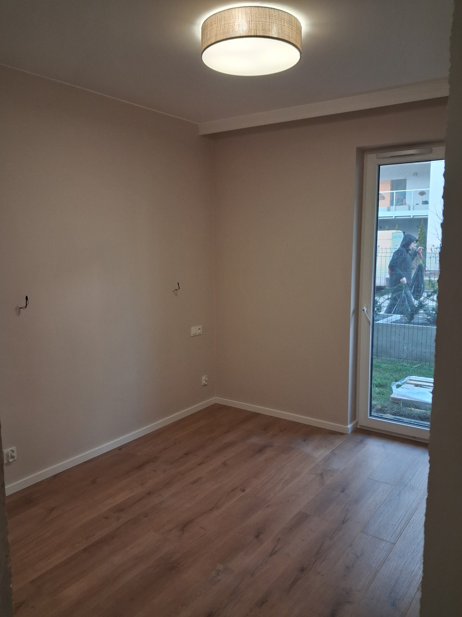 Pusty pokój po malowaniu ścian na beżowo i ułożeniu paneli podłogowych z widocznym oknem balkonowym i lampą sufitową.