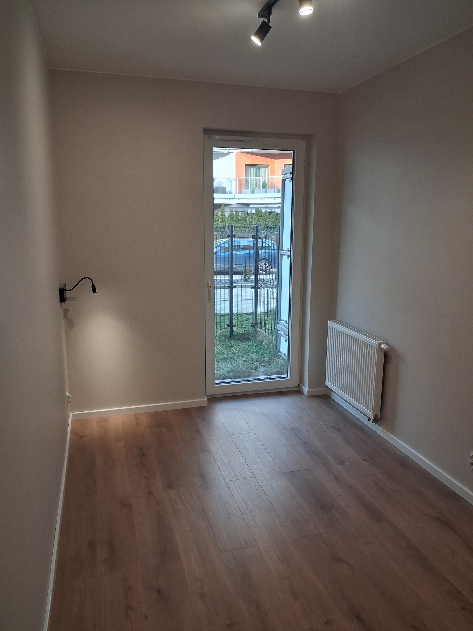 Pusty pokój z panelami podłogowymi w odcieniach brązu, świeżo pomalowanymi ścianami w kolorze beżowym, grzejnikiem i lampą ścienną, widok na ogród przez przeszklone drzwi balkonowe.