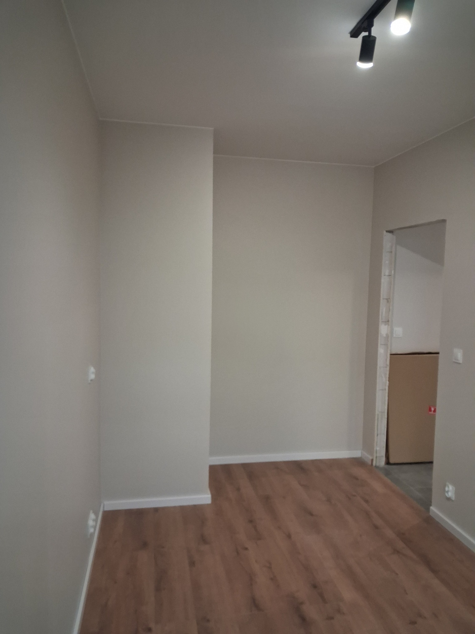 Pusty pokój po remoncie z panelami podłogowymi w odcieniach brązu, świeżo pomalowane ściany w kolorze jasnego beżu i nowoczesne oświetlenie sufitowe na szynie. W tle widoczne drzwi do innego...