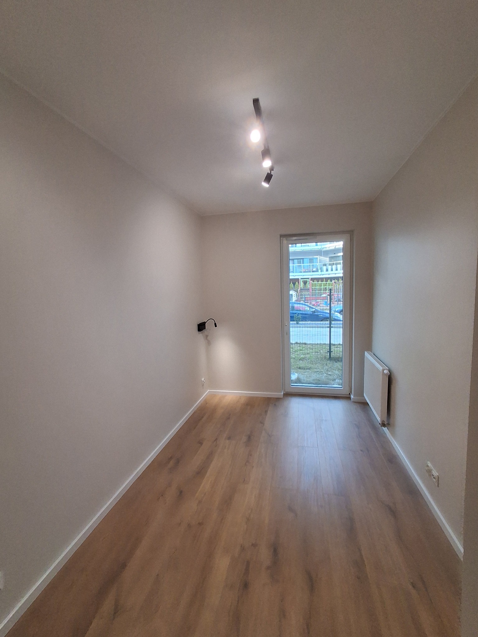 Pusty pokój w Łodzi po remoncie, z drewnianą podłogą, świeżo pomalowanymi ścianami w odcieniach beżu, oświetleniem sufitowym punktowym i grzejnikiem przy balkonie.