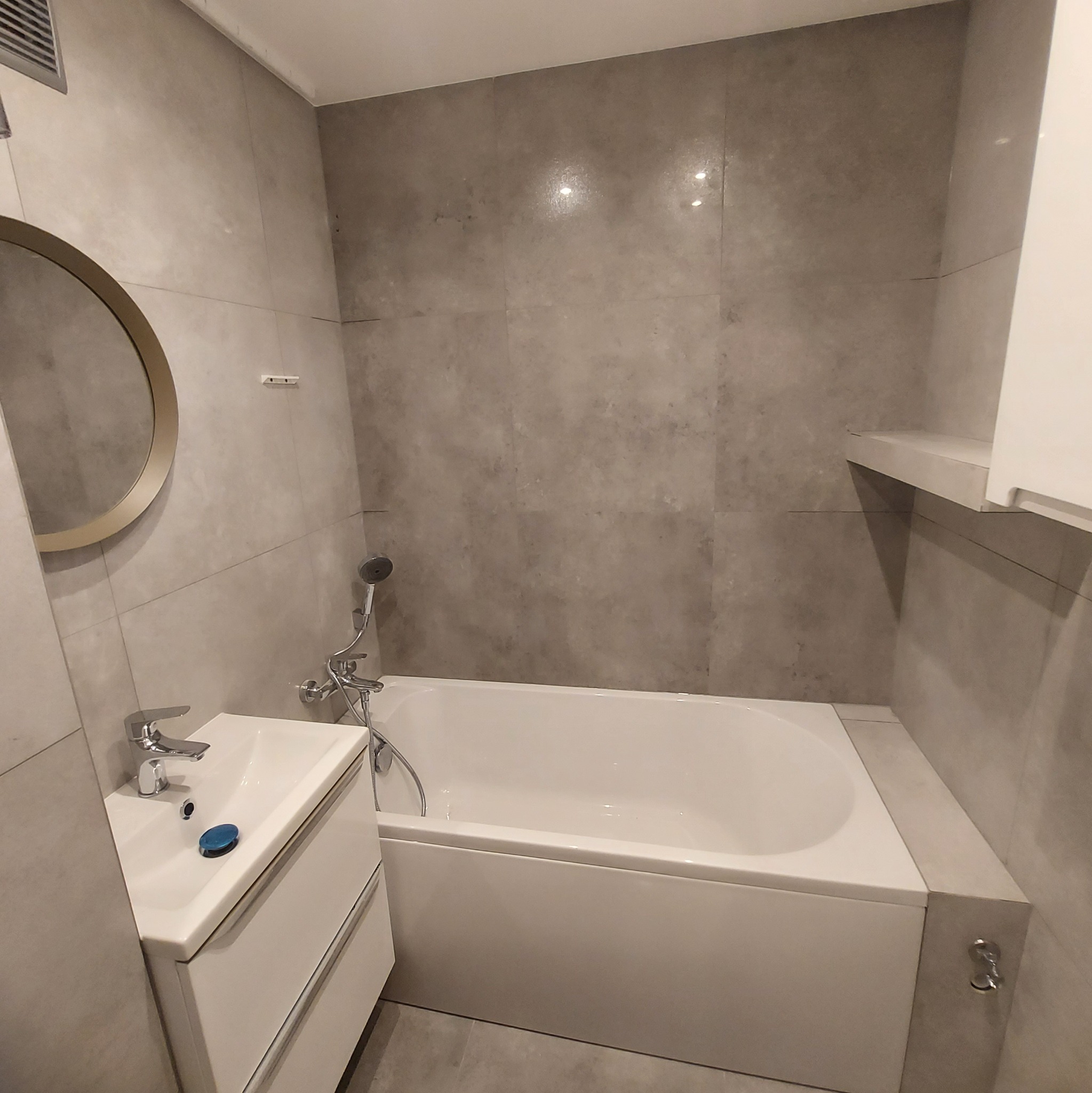 Nowoczesna łazienka z szarymi płytkami, owalnym lustrem w złotej ramie, białą umywalką z szafką i wanną z zamontowaną słuchawką prysznicową.