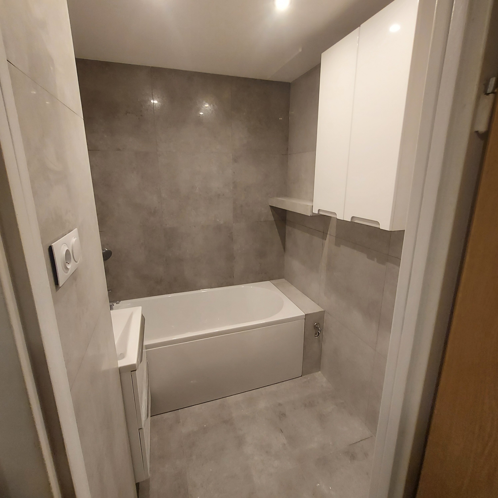 Nowoczesna łazienka z szarymi płytkami imitującymi beton, białą wanną, szafką pod umywalką i wiszącymi szafkami. Na ścianie widoczny biały włącznik światła.