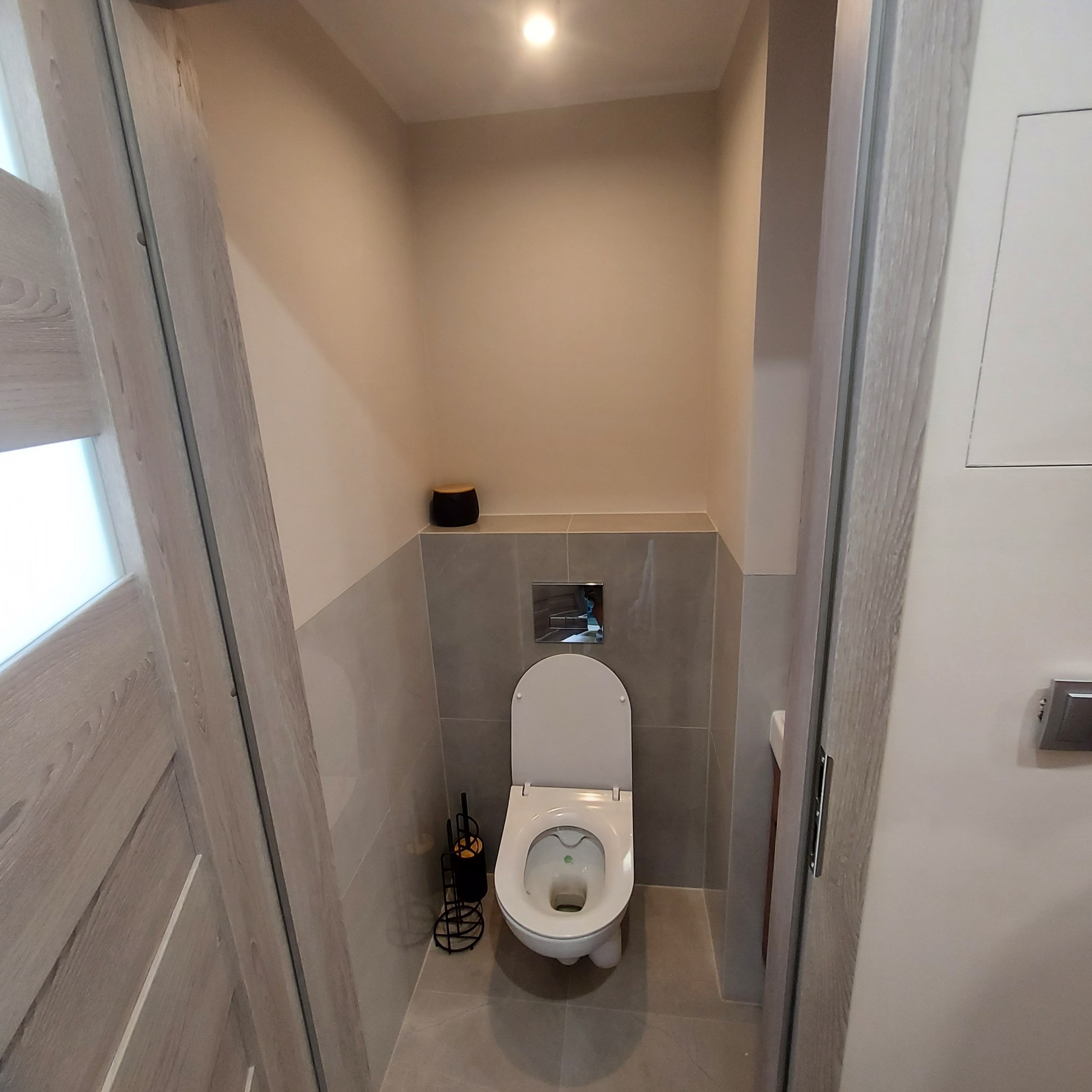 Niewielka toaleta z szarymi płytkami i wiszącą muszlą klozetową, widok zza uchylonych drzwi o drewnianej fakturze, czarny pojemnik na szczotkę toaletową w rogu.