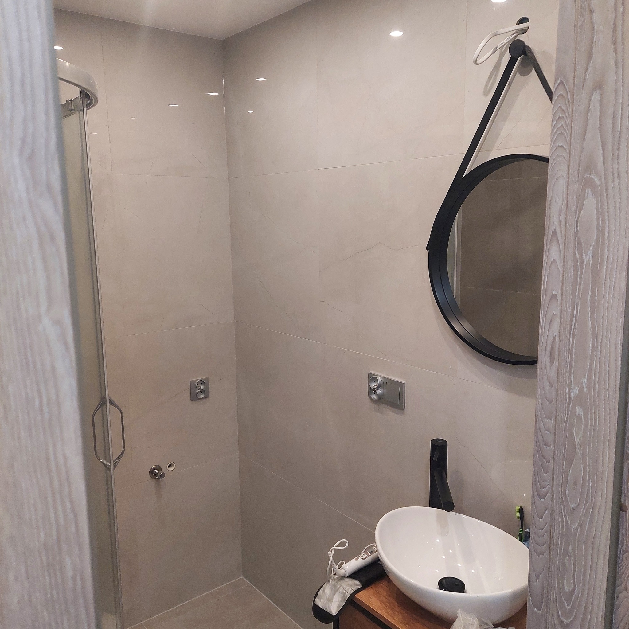 Nowoczesna łazienka z kabiną prysznicową, umywalką nablatową na drewnianym blacie, okrągłym lustrem w czarnej ramie i szarymi płytkami na ścianach. Na blacie leży suszarka do włosów.