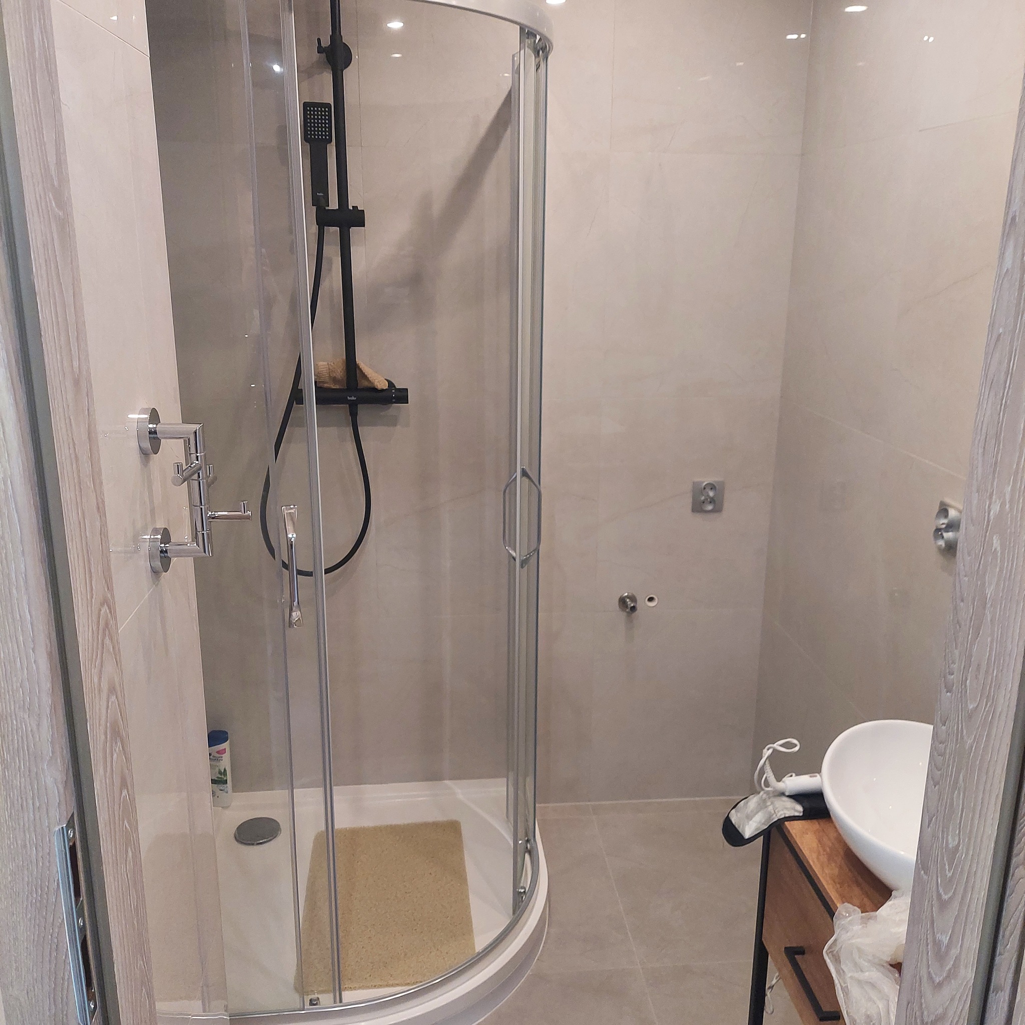 Nowoczesna kabina prysznicowa z czarną armaturą i beżowymi płytkami w łazience, widoczna umywalka nablatowa i haczyki na ścianie, całość w jasnej tonacji.