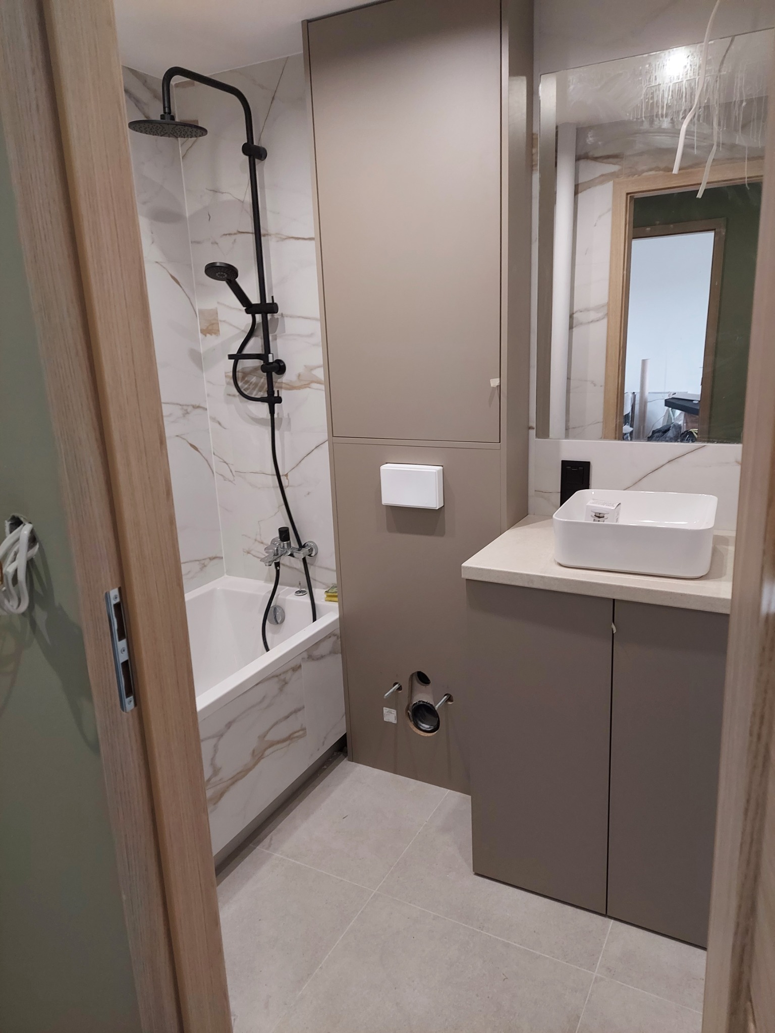 Nowoczesna łazienka z beżowymi szafkami, białą umywalką nablatową i baterią, kabiną prysznicową typu walk-in z czarną armaturą oraz płytkami imitującymi marmur na ścianach i podłodze.